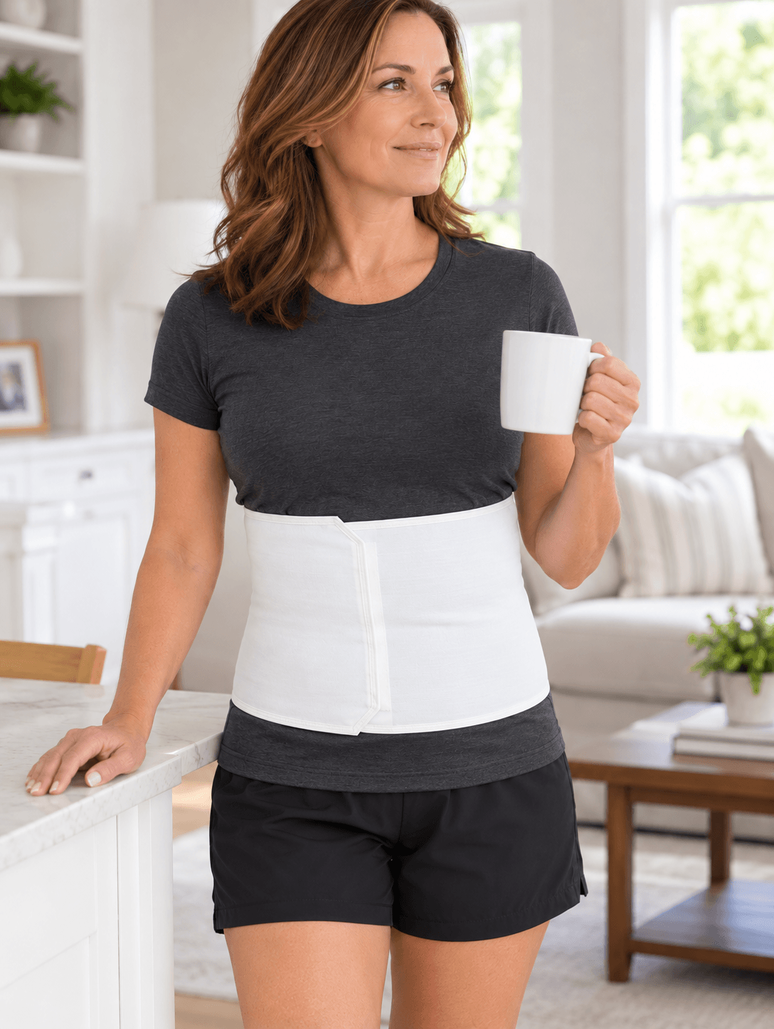 Woman wearing a white EzyWrap abdominal support binder over a charcoal shirt while standing at home with a coffee mug.